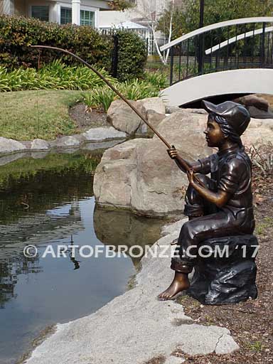 Gone Fishing bronze sculpture of girl sitting on rock fishing. Wearing overalls and baseball hat.