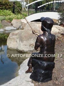 Gone Fishing bronze sculpture of girl sitting on rock fishing. Wearing overalls and baseball hat.